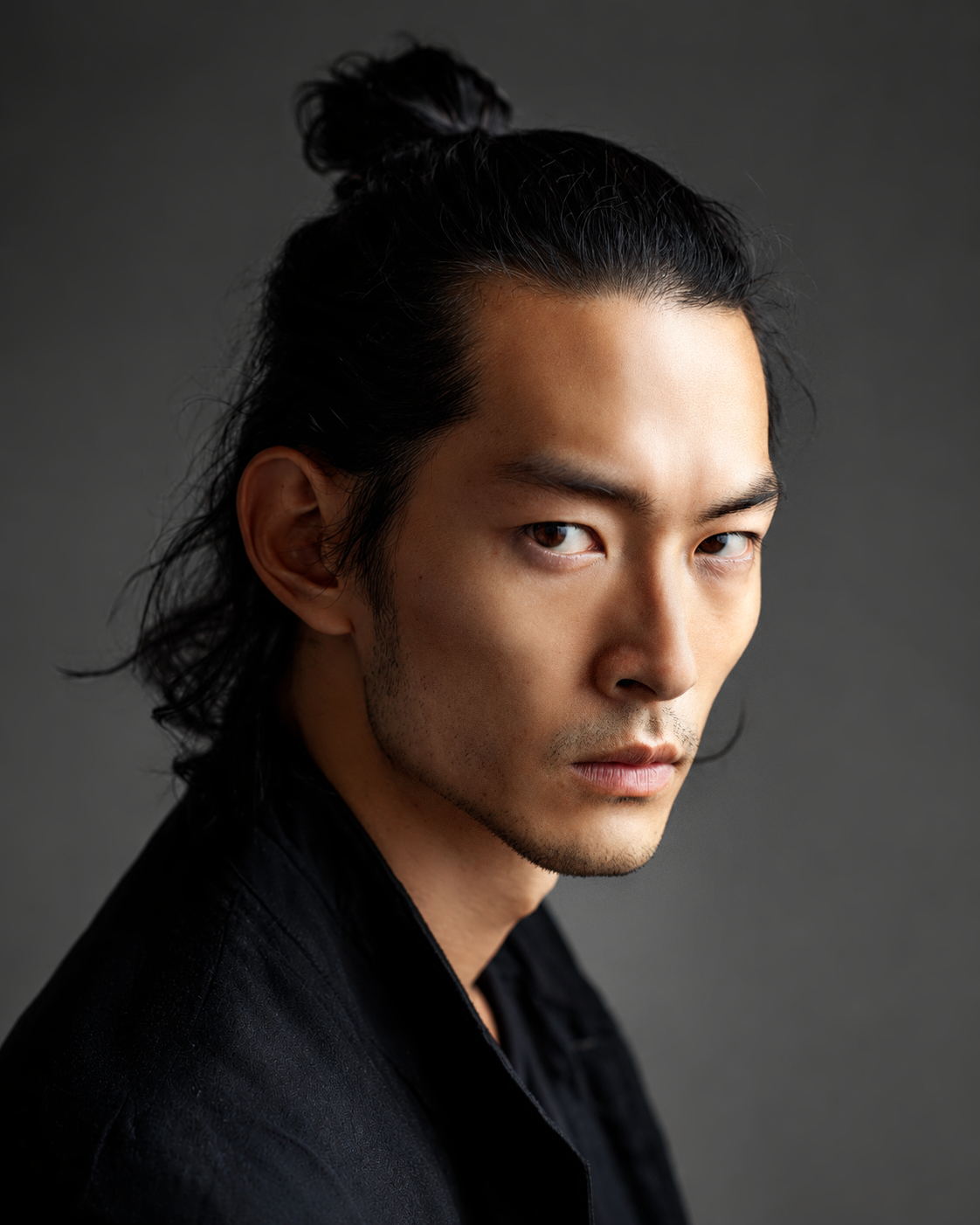Hiro, male virtual AI fashion model with dark hair in a topknot and subtle facial hair, dramatic close-up portrait in black clothing by VIDA AI